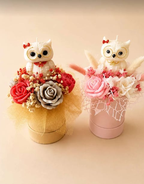Two decorative owl figurines with red bows sitting atop flower arrangements in gold and pink cylindrical boxes