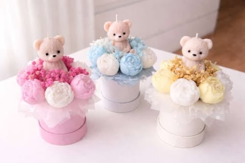 Three decorative flower arrangements with teddy bears in pink, blue, and yellow colors displayed on a white surface