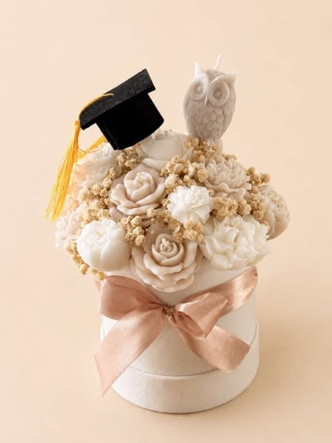 Decorative floral arrangement with cream and beige roses, a white owl figurine, graduation cap with gold tassel, and pale pink bow in a white pot
