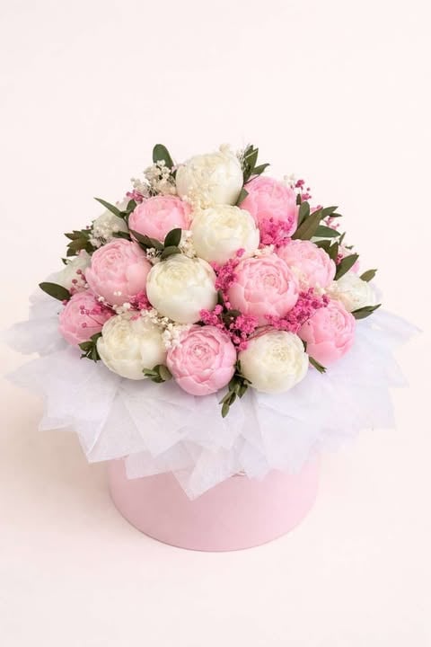 Bouquet of pink and white peonies with green foliage in a pink round box with white ribbon wrap