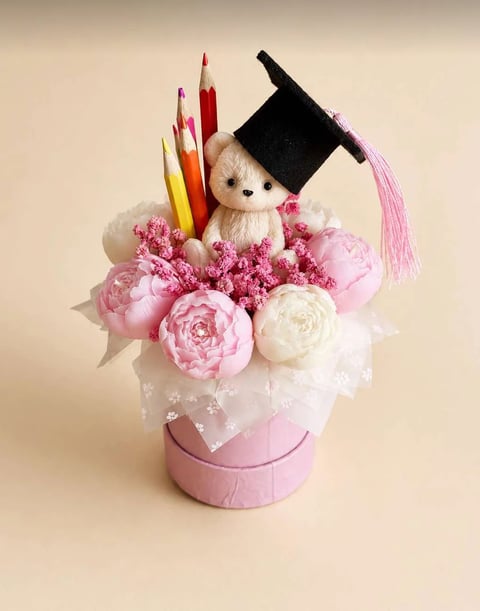Decorative arrangement in pink box with teddy bear wearing graduation cap, pink and white peonies, colored pencils, and tassel